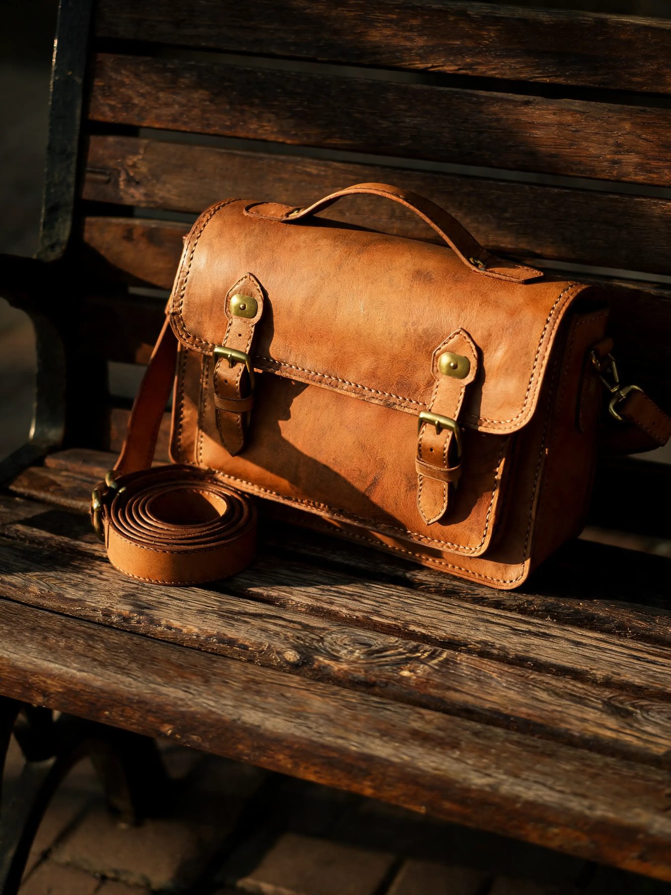 Vegetable-tanned leather satchel with brass buckles beside a coiled belt on a wooden bench
